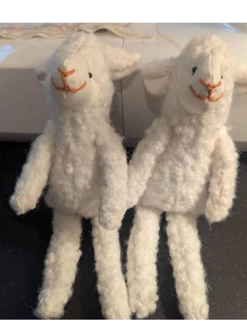 May include: Two handmade white lamb plush toys. Each lamb has a fluffy, textured body, small black eyes, and an orange stitched mouth and nose. The lambs are standing upright with their arms and legs extended.