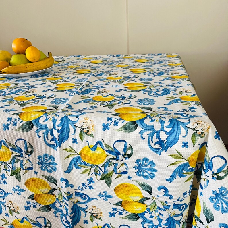 May include: White tablecloth with a repeating pattern of yellow lemons, blue floral accents, and green leaves. The tablecloth is covering a table and has a fruit bowl with bananas and other fruits.