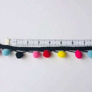 Könnte beinhalten: Schwarzer Besatz mit bunten Bommeln. Der Besatz ist 17,8 cm lang und hat blaue, schwarze, rosa, rote, gelbe und rosa Bommeln.