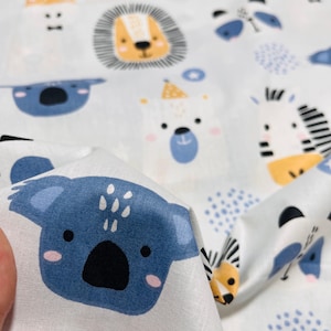 May include: A white fabric featuring a repeating pattern of cartoon animal faces including blue koalas, yellow giraffes, orange-maned lions, and black-and-white striped zebras, interspersed with small blue polka dot clusters and star shapes.