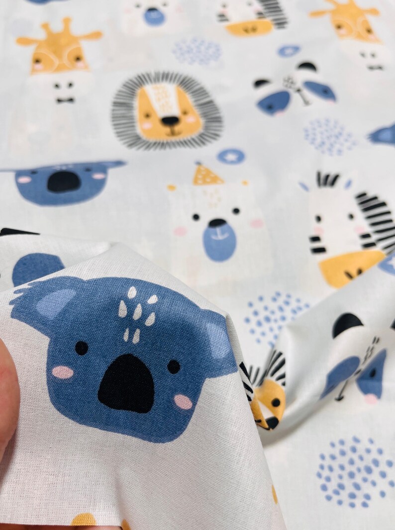 May include: A white fabric featuring a repeating pattern of cartoon animal faces including blue koalas, yellow giraffes, orange-maned lions, and black-and-white striped zebras, interspersed with small blue polka dot clusters and star shapes.