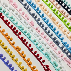 Könnte beinhalten: Sortiment an bunten Pompon-Zierbändern. Das Band besteht aus einem dünnen weißen Band mit Pompons in verschiedenen Farben, darunter Rot, Orange, Gelb, Grün, Blau, Lila und Pink.