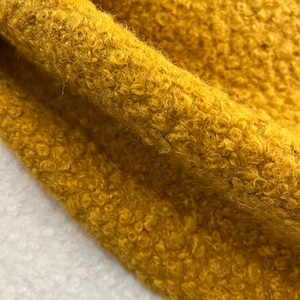 Może przedstawiać: Zbliżenie na miękką, puszystą tkaninę w ciepłym, musztardowym kolorze. Tkanina ma fakturowaną, kręconą powierzchnię.