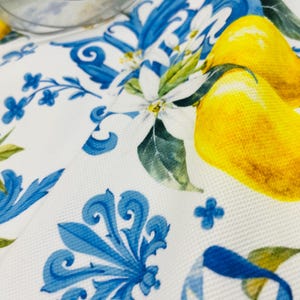 May include: White fabric with a blue floral design and yellow lemons. The fabric features a repeating pattern of blue scrollwork, small blue flowers, and yellow lemons with green leaves. A clear glass bowl is in the background.