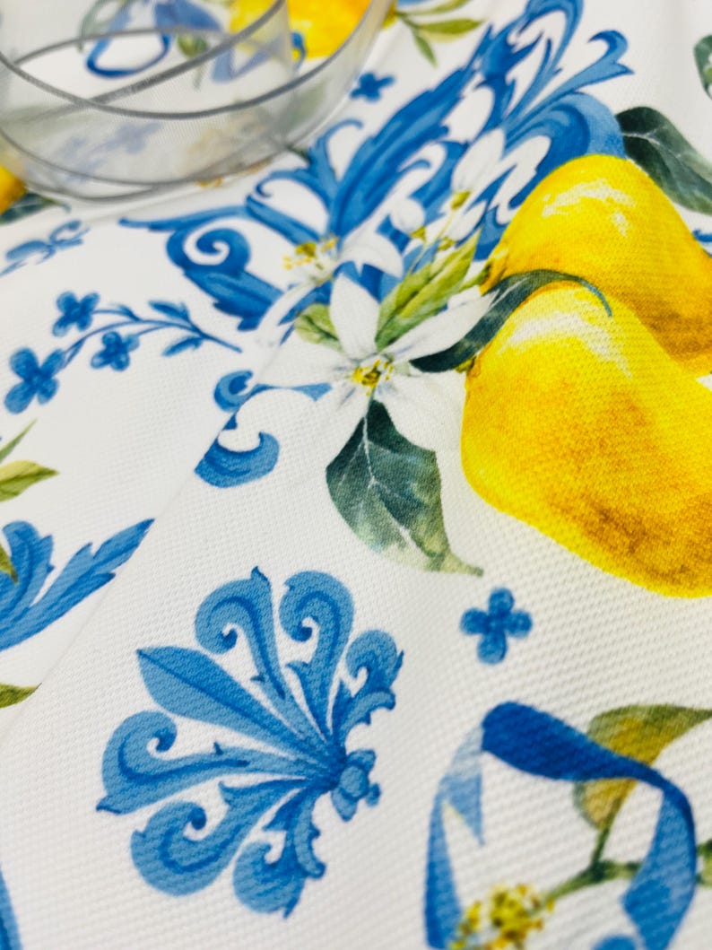 May include: White fabric with a blue floral design and yellow lemons. The fabric features a repeating pattern of blue scrollwork, small blue flowers, and yellow lemons with green leaves. A clear glass bowl is in the background.