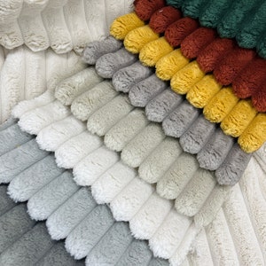 May include: Close-up of a multi-colored plush fabric with a ribbed texture. The colors include shades of white, gray, yellow, brown, and green. The fabric has a soft, fuzzy appearance, and the ribbed pattern creates a visually interesting design.