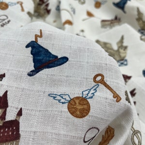 White fabric with a repeating pattern of blue wizard hats, gold keys, brown flying snitches, and brown castles. The fabric is textured and has a subtle sheen.
