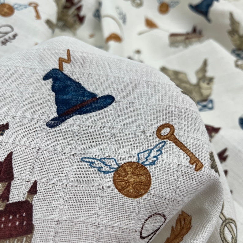 White fabric with a repeating pattern of blue wizard hats, gold keys, brown flying snitches, and brown castles. The fabric is textured and has a subtle sheen.