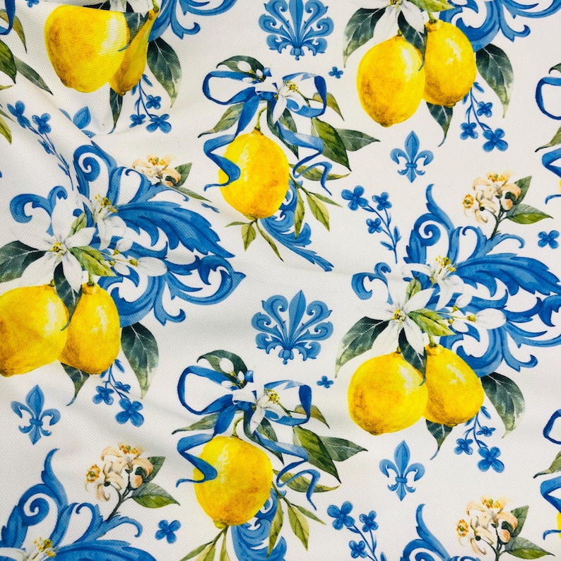 May include: White fabric with a repeating pattern of yellow lemons, blue ribbons, and floral accents. The design includes blue scrollwork, white flowers, and green leaves. The lemons are bright yellow.