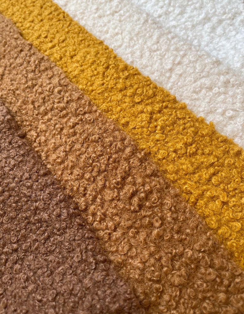 May include: Close-up of a fabric sample with a textured, curly pile. The fabric is divided into diagonal stripes of varying colours, including brown, tan, mustard yellow, and off-white. The texture appears soft and plush.