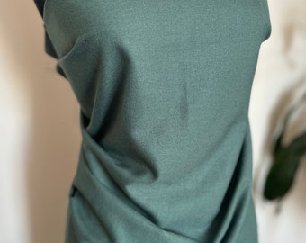 Puede incluir: Una tela verde oscuro drapeada sobre un maniquí negro. Un collar dorado con un diseño floral y un colgante en forma de lágrima se muestra en el maniquí. La tela tiene una textura sutil.