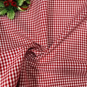 May include: Red and white checkered fabric, a classic gingham pattern, is displayed in a close-up shot. The fabric is draped, showing folds and texture. A small decorative floral element with red berries and green leaves is in the upper left corner.