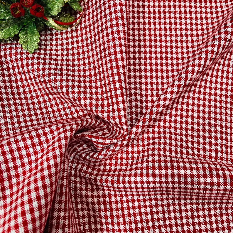 May include: Red and white checkered fabric, a classic gingham pattern, is displayed in a close-up shot. The fabric is draped, showing folds and texture. A small decorative floral element with red berries and green leaves is in the upper left corner.