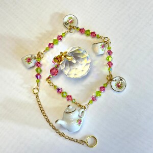 May include: A delicate charm bracelet featuring a crystal ball, a teapot, and several miniature teacups with floral designs. The bracelet is made with pink and green crystal beads and a gold chain.