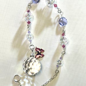 Puede incluir: Un atrapasoles decorativo de cristal con una esfera facetada, cuentas de vidrio transparentes e iridiscentes y detalles en morado y rosa. Un peque&ntilde;o dije de mariposa est&aacute; adjunto. El atrapasoles est&aacute; suspendido por una cadena plateada.