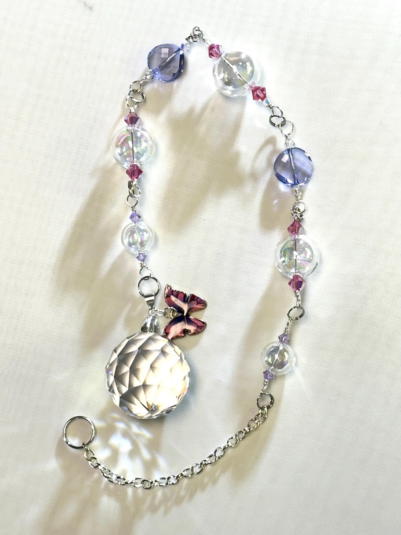 Puede incluir: Un atrapasoles decorativo de cristal con una esfera facetada, cuentas de vidrio transparentes e iridiscentes y detalles en morado y rosa. Un peque&ntilde;o dije de mariposa est&aacute; adjunto. El atrapasoles est&aacute; suspendido por una cadena plateada.