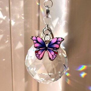 Puede incluir: Un atrapasoles de bola de cristal transparente con un dije de mariposa decorativo. La mariposa tiene alas rosas y moradas con detalles rojos y un marco de metal plateado. El atrapasoles cuelga de una cadena plateada.