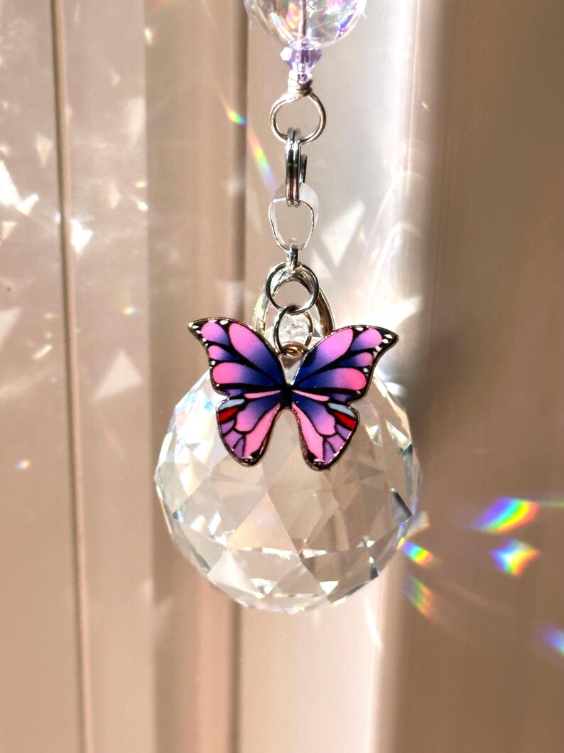 Puede incluir: Un atrapasoles de bola de cristal transparente con un dije de mariposa decorativo. La mariposa tiene alas rosas y moradas con detalles rojos y un marco de metal plateado. El atrapasoles cuelga de una cadena plateada.