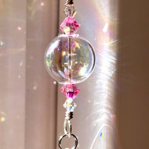Puede incluir: Un globo de vidrio transparente e iridiscente con detalles de cristal rosa y transparente, suspendido por una cadena plateada. El globo refleja la luz, creando un efecto arco&iacute;ris. Pieza decorativa para el hogar.