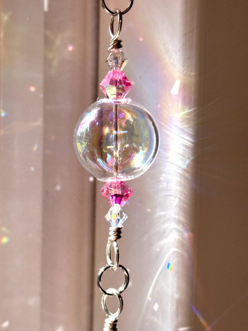 Puede incluir: Un globo de vidrio transparente e iridiscente con detalles de cristal rosa y transparente, suspendido por una cadena plateada. El globo refleja la luz, creando un efecto arco&iacute;ris. Pieza decorativa para el hogar.