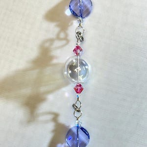 Puede incluir: Un adorno de cuentas con una cuenta redonda transparente, dos cuentas redondas azules y cuentas de cristal rosa. Las cuentas est&aacute;n conectadas por anillos y alambre de metal plateado. El adorno cuelga sobre un fondo blanco.