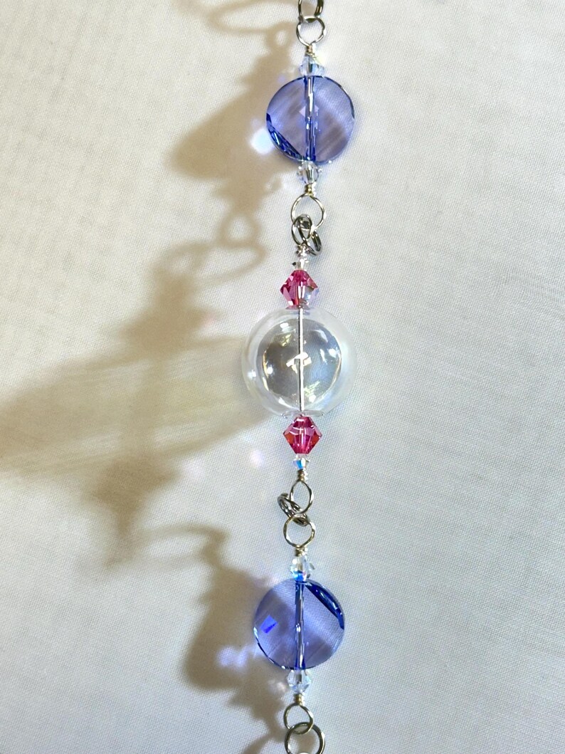 Puede incluir: Un adorno de cuentas con una cuenta redonda transparente, dos cuentas redondas azules y cuentas de cristal rosa. Las cuentas est&aacute;n conectadas por anillos y alambre de metal plateado. El adorno cuelga sobre un fondo blanco.