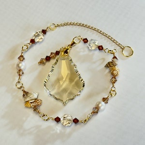 以下が含まれることがあります： 透明なクリスタル製の涙型のペンダントと茶色と白のビーズが付いた金色のチェーン。チェーンには小さな貝殻と真珠も付いています。