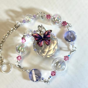 Puede incluir: Un atrapasoles decorativo con una bola de cristal facetada y un dije de mariposa. El atrapasoles est&aacute; adornado con cuentas transparentes, rosas y moradas y orbes de vidrio transparente, conectados por una cadena plateada. El dije de mariposa tiene alas rosas y moradas.