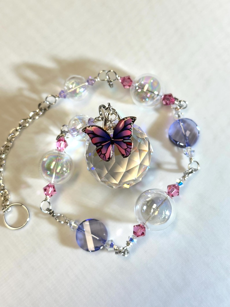 Puede incluir: Un atrapasoles decorativo con una bola de cristal facetada y un dije de mariposa. El atrapasoles est&aacute; adornado con cuentas transparentes, rosas y moradas y orbes de vidrio transparente, conectados por una cadena plateada. El dije de mariposa tiene alas rosas y moradas.