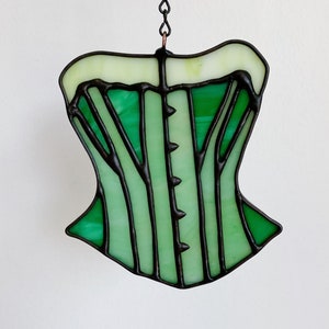 May include: A green stained glass corset with black metal framing. The corset is hanging from a chain.