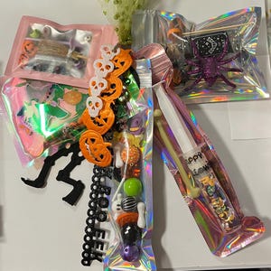 May include: A collection of Halloween-themed craft supplies including holographic pouches filled with assorted beads, charms, and miniature decorations. Items include orange pumpkin charms, white skull links, a black 'BOO' sign, and a syringe filled with confetti.