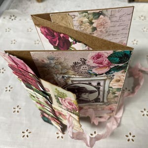 Pu&ograve; includere: Un album di ritagli rosa e marrone con un design floreale e una foto di una donna con un cappello. L'album &egrave; decorato con nastri e ha un design a strati.