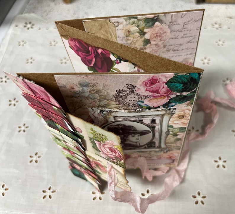 Pu&ograve; includere: Un album di ritagli rosa e marrone con un design floreale e una foto di una donna con un cappello. L'album &egrave; decorato con nastri e ha un design a strati.