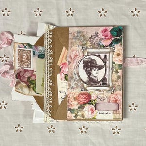 Pu&ograve; includere: Un diario di carta marrone con un bordo di pizzo e un nastro rosa. La copertina &egrave; decorata con motivi floreali, un ritratto vintage di una donna e il testo "Austr.alia".