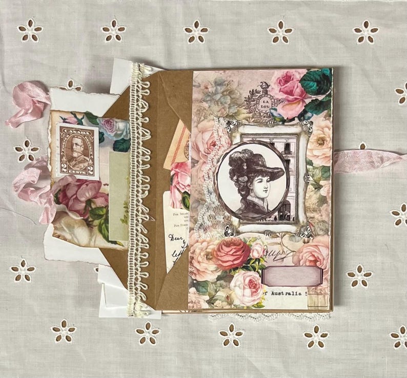 Pu&ograve; includere: Un diario di carta marrone con un bordo di pizzo e un nastro rosa. La copertina &egrave; decorata con motivi floreali, un ritratto vintage di una donna e il testo "Austr.alia".