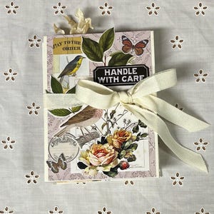 Könnte beinhalten: Ein rosa-weißes Journal mit floralem und Vogel-Design. Das Journal ist mit einem weißen Band gebunden und hat ein "Handle with Care"-Etikett. Das Journal liegt auf einem weißen Hintergrund mit einem floralen Muster.