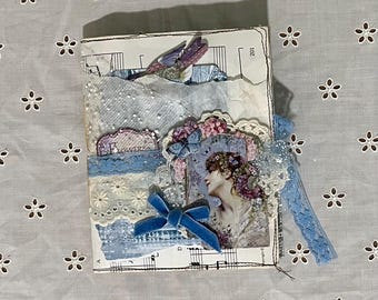 Può includere: Un diario fatto a mano con un'estetica vintage. La copertina presenta un collage di pizzo, spartiti musicali e un ritratto di donna con fiori. Impreziosito da un fiocco di velluto blu e un nastro. La parola "FLOWERS" è visibile.