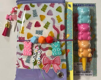 Può includere: Un astuccio trasparente con motivo a caramelle gommose, cerniera viola, ciondolo a nappa rosa e catenella di perline a forma di orsetto. Include una toppa a fiocco rosa e una penna impilabile a forma di orsetto blu, rosa e arancione su blister.