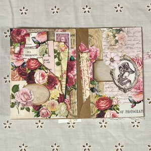 Pu&ograve; includere: Un album di scrapbooking a tema floreale. La copertina &egrave; decorata con rose rosa e rosse, foglie e altri elementi floreali. L'album &egrave; aperto per rivelare una pagina con una foto in stile vintage di una donna con un cappello. La pagina contiene anche una nota scritta a mano che dice "Of Photography".
