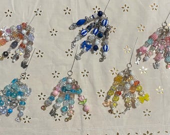 Può includere: Una collezione di ciondoli di perline in vari colori, tra cui blu, rosa, giallo e trasparente. Ogni ciondolo presenta una disposizione unica di perline, alcune con dettagli a farfalla e fiocco. I ciondoli sono attaccati a spilli di metallo.