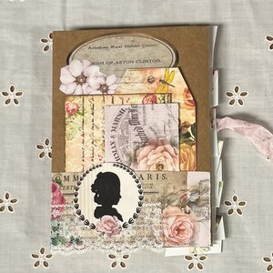 Pu&ograve; includere: Un album di scrapbooking in stile vintage con una copertina in carta marrone e vari abbellimenti in carta, tra cui motivi floreali, una silhouette di una donna e una libellula. L'album &egrave; decorato con pizzo e ha un nastro rosa legato intorno.
