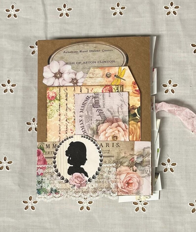 Pu&ograve; includere: Un album di scrapbooking in stile vintage con una copertina in carta marrone e vari abbellimenti in carta, tra cui motivi floreali, una silhouette di una donna e una libellula. L'album &egrave; decorato con pizzo e ha un nastro rosa legato intorno.