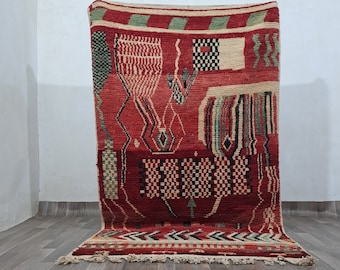 May include: A red, green, and cream colored rug with a geometric pattern. The rug is folded in half and leaning against a white wall.