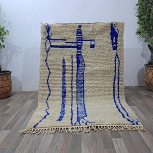 Può includere: Un tappeto marocchino bianco e blu con un motivo geometrico. Il tappeto ha un bordo a frangia ed è fatto di lana.