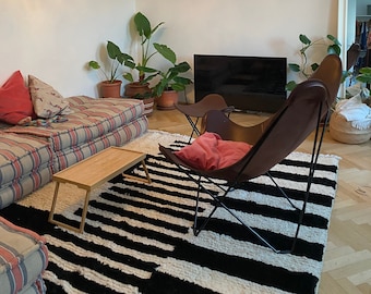 May include: A living room with a black and white striped rug, a brown leather butterfly chair, a couch with red and white striped cushions, a wooden coffee table, and a chandelier hanging from the ceiling.