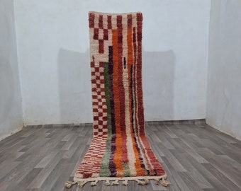 May include: A long, rectangular rug with a geometric pattern in shades of red, orange, green, and white. The rug has a fringe on both ends and is lying on a wooden floor.