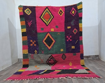 May include: A vibrant, hand-woven rug with a geometric design. The rug features a bold color palette of pink, green, orange, and purple, with diamond and square patterns. The rug is displayed against a white wall and wooden floor.