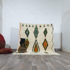 May include: Cream-colored, hand-woven rug with a diamond pattern in green, orange, and black. The rug has a fringed edge and a geometric design. The rug is placed on a wooden floor.