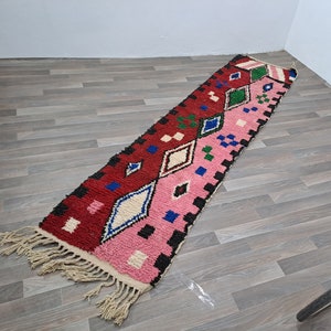 Peut inclure: Un long tapis rectangulaire au design divisé, avec une moitié rouge foncé et l'autre rose clair. Le tapis est orné de motifs géométriques en losange blancs, bleus et verts, agrémentés de carrés noirs et de franges de couleur crème aux extrémités.