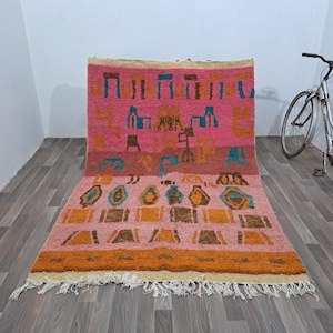 May include: A pink, orange, and blue Moroccan rug with a geometric pattern. The rug has a fringe border and is made of wool.